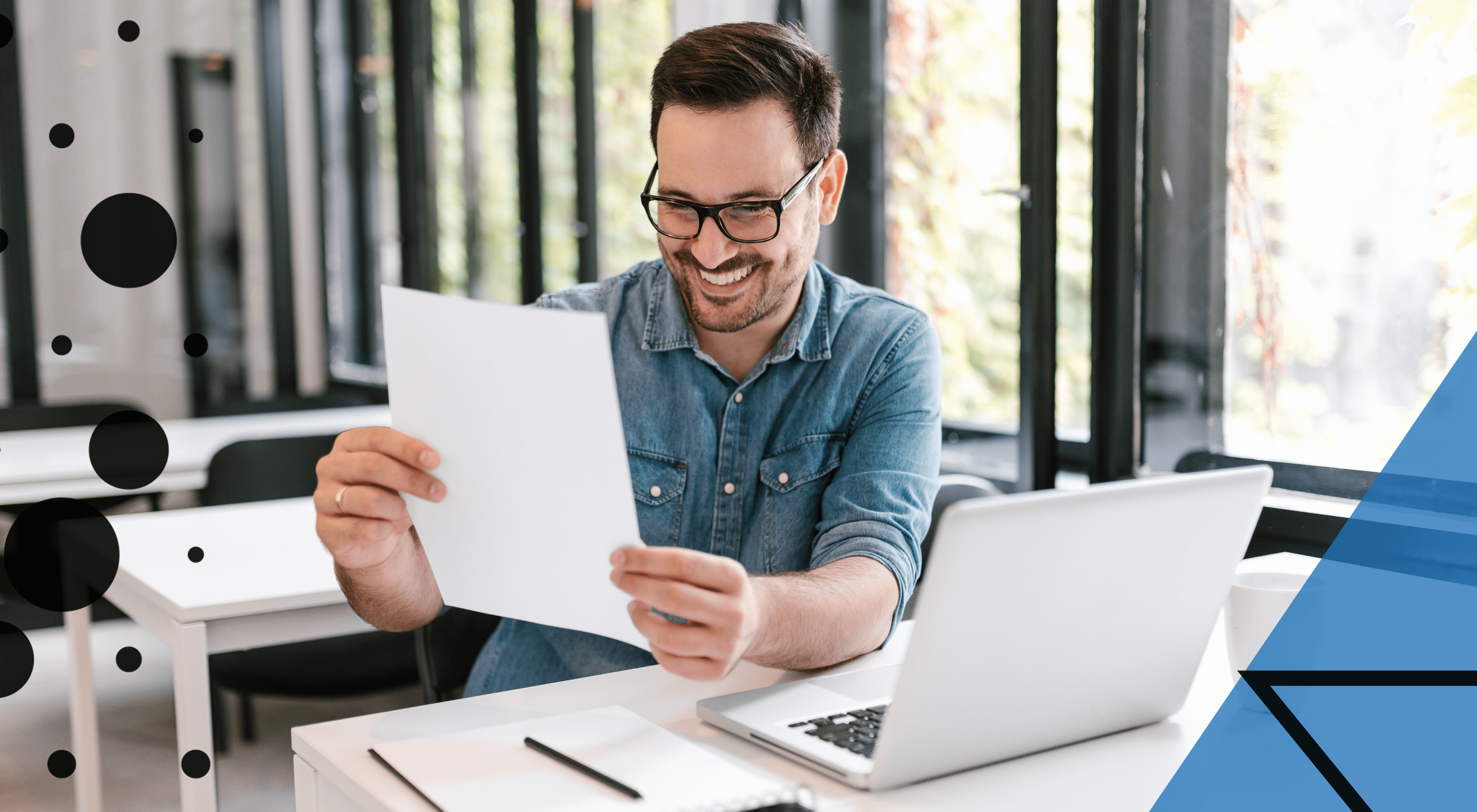 Man at desk on laptop looking pleased as he looks at his results on paper