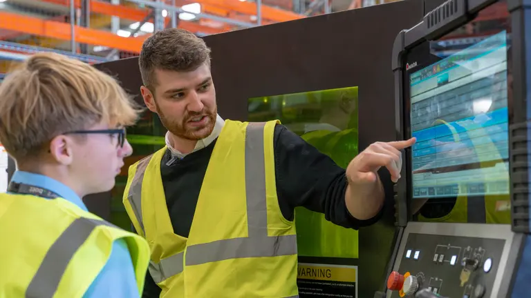 NMD Two men in safety vests standing in front of a large monitor.