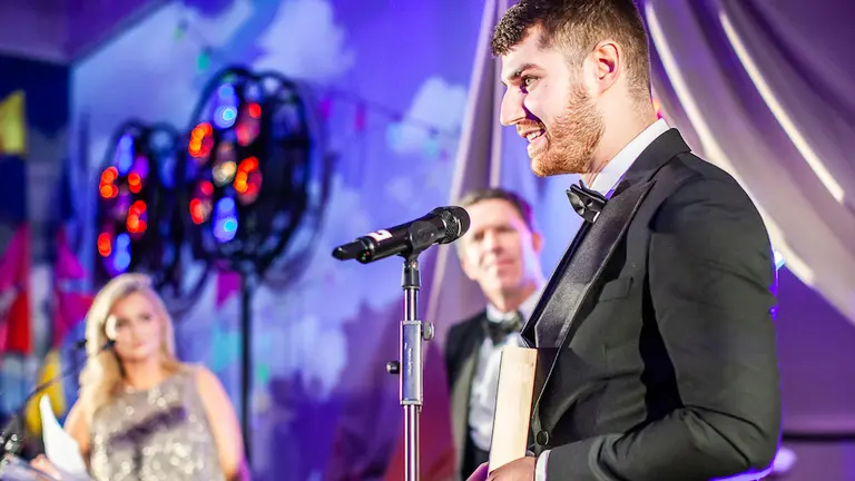 Man with make uk award in black suit standing on stage in front of microphone.