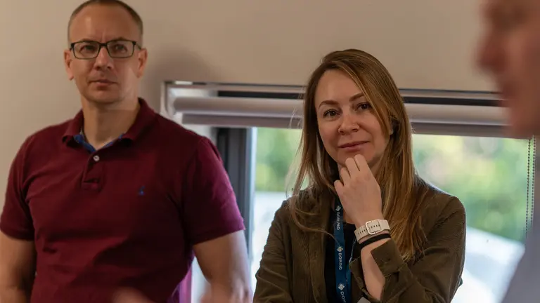 A man in a maroon polo shirt and a woman in a brown jacket face each other with a third person.