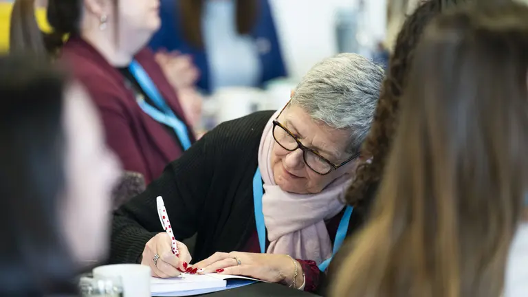 A woman wearing glasses and scarf is writing on a notepad at a conference.