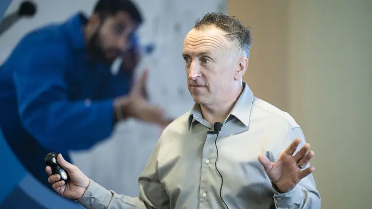 Man gesturing with one hand, holding a remote in the other, speaking to an audience.