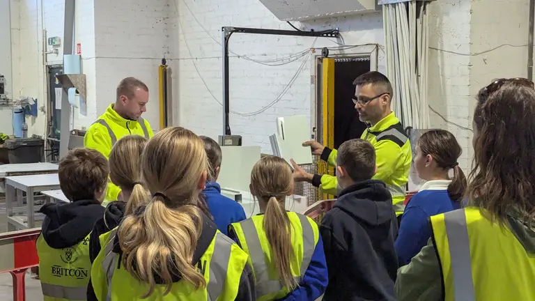 Children wearing yellow vests listen to a man explain something in a factory on national manufacturing day.