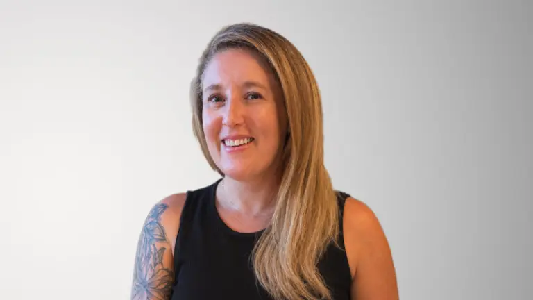 Blonde woman with tattoos on arms smiling and wearing black sleeveless top.