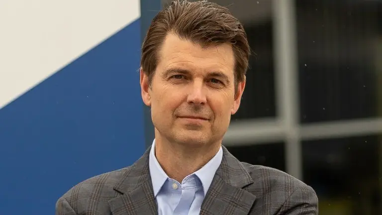 Man in a suit smiling for the camera with folded arms standing in front of a blue wall.