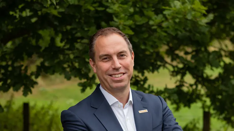 Man in suit stands with arms folded outdoors with a smile.
