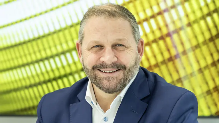Man in blue jacket, white shirt, and beard smiling in front of yellow background.