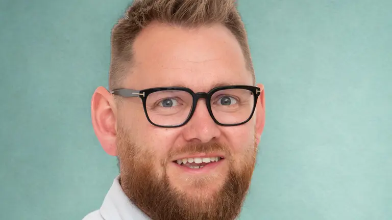 A man wearing glasses and a white shirt smiles against a teal background