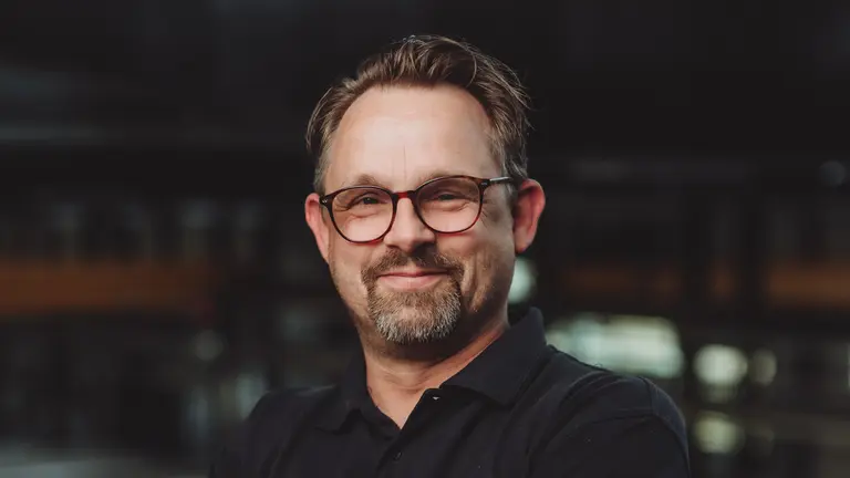 A smiling man wearing glasses and a black shirt with a logo on it.