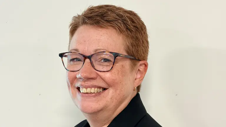 Smiling woman wearing glasses and a black jacket, standing in front of a white wall.