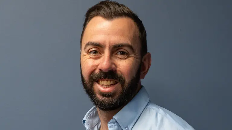 A man with a beard smiles at the camera, wearing a light blue shirt with MGS logo.
