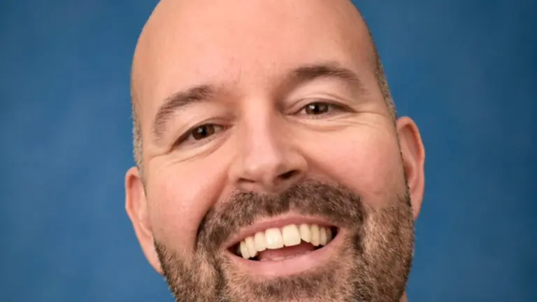 A man with a beard smiles and shows his teeth against a blue background.