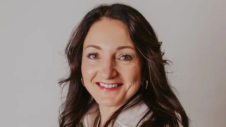 A smiling woman in a white jacket poses with arms crossed.