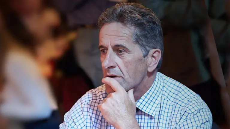 A man in a blue and white checkered shirt sits in a crowd, hand on his chin.