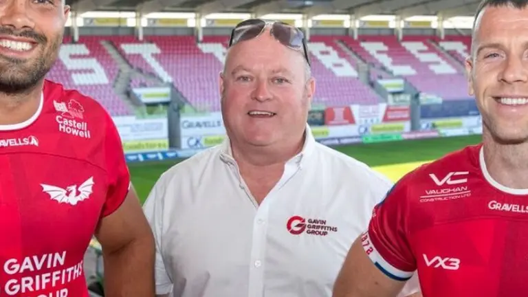 Three men stand in front of an empty stadium. Two wear red jerseys and one wears a white shirt.