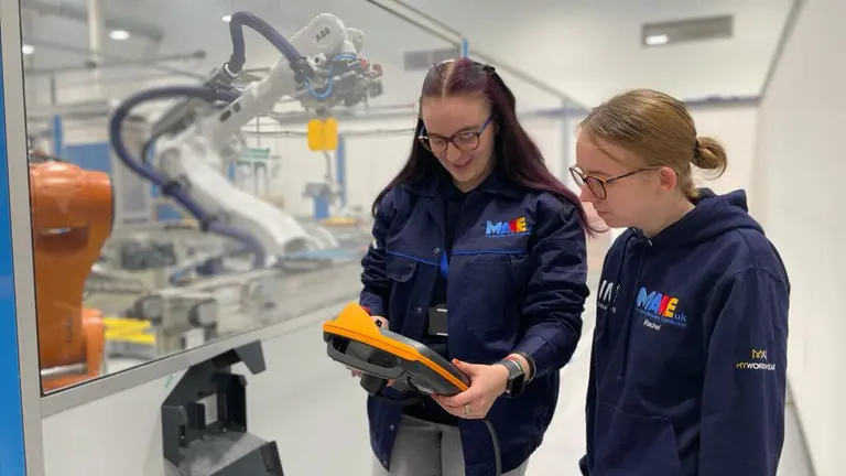 Two women in blue jackets review a tablet in a factory with an arm-like machine behind them.