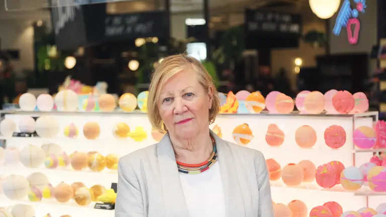 A woman in a gray blazer stands in front of a colorful display of bath bombs.