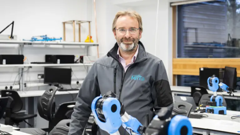A man stands in a workshop with robotic arms in front of him and smiling for the camera.