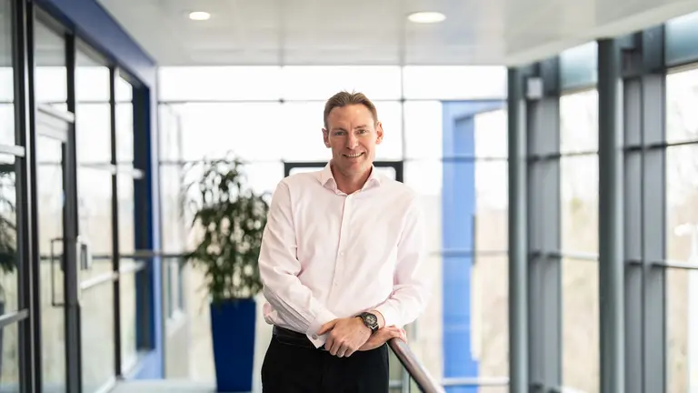 A man in a pink shirt and black pants stands in a modern office building, smiling for the camera.