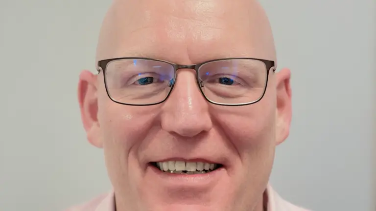Man wearing glasses smiling for the camera wearing a pink shirt.