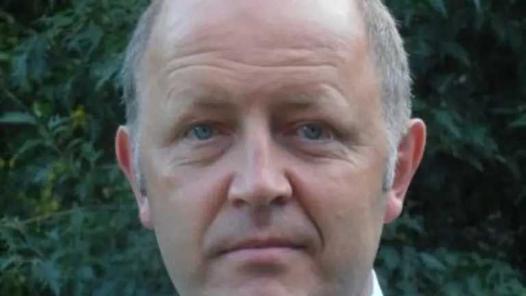 A man in a blue and white checkered shirt with a red tie looks into the camera.