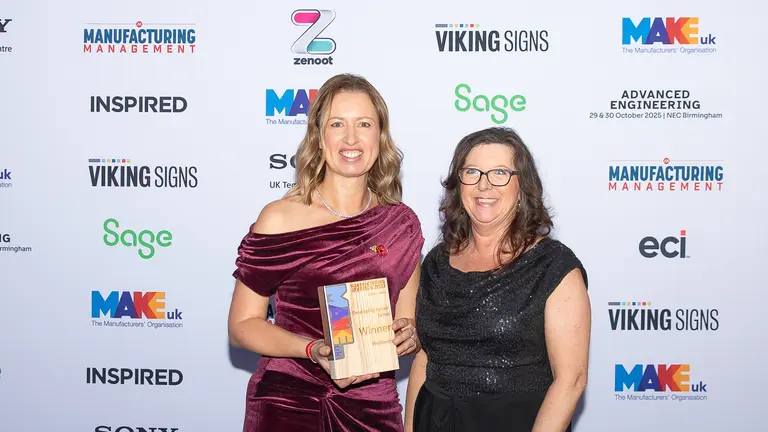 Two women stand with an award, one holding it, in front of a backdrop with logos.