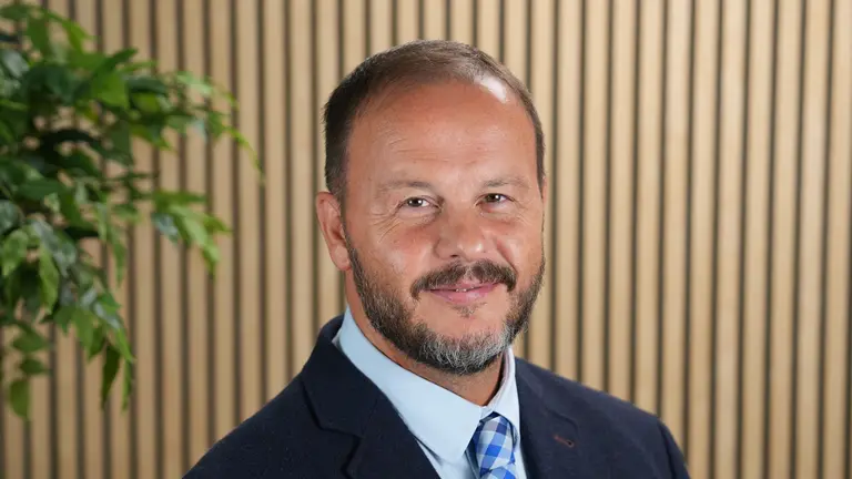 Man with beard in suit and tie smiling standing beside a plant.