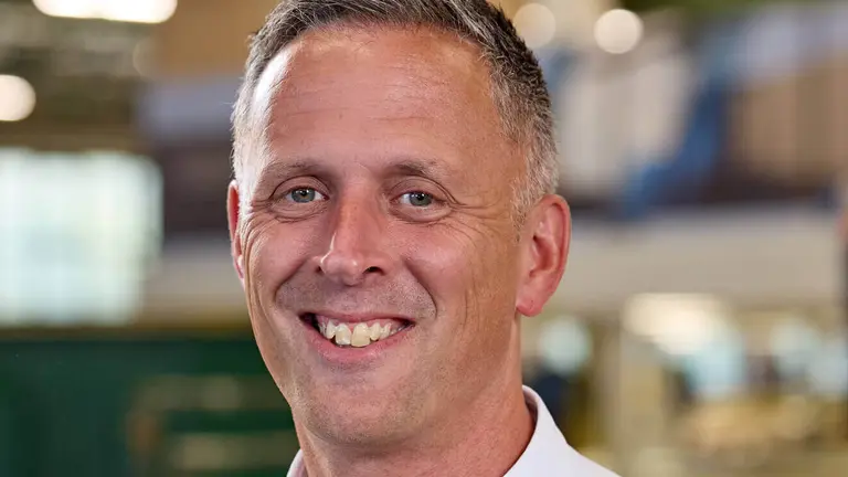 Man wearing white shirt with company logo on left chest smiling in a factory.