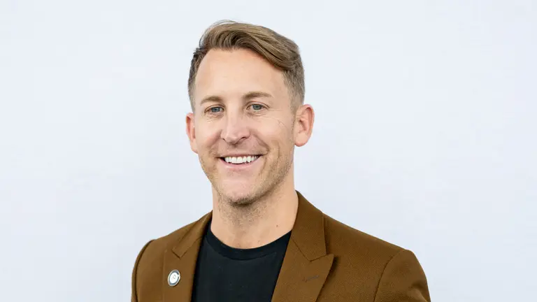 Man in brown blazer, black shirt, and pin, smiling at camera.