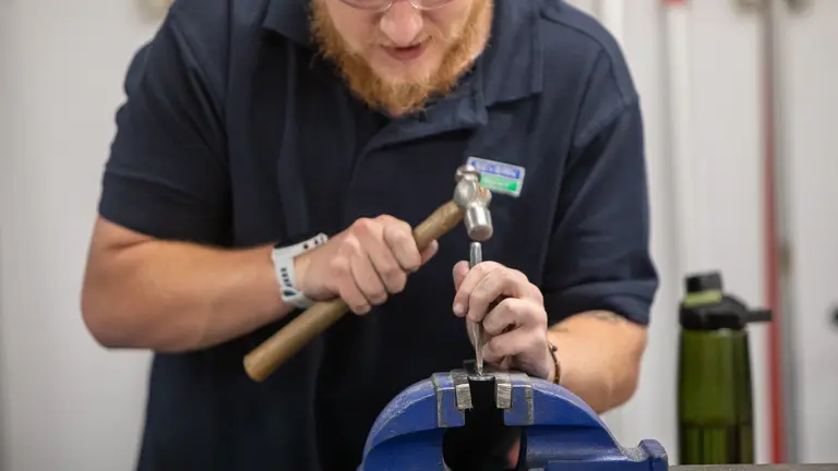 Apprentice with beard hammering tool into metal object in blue vise grip.