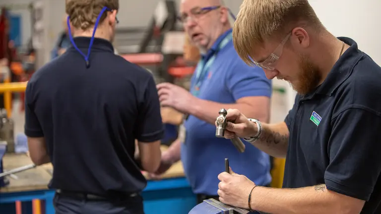 Three men in a workshop with the man in the middle using a tool.