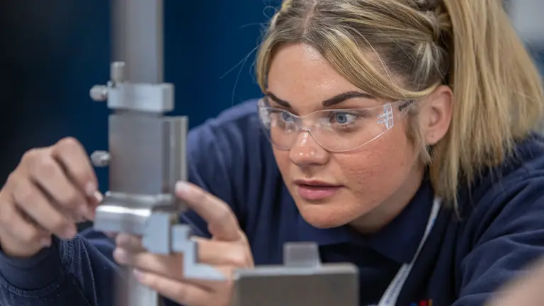 An apprentice wearing safety goggles examines a metal object.