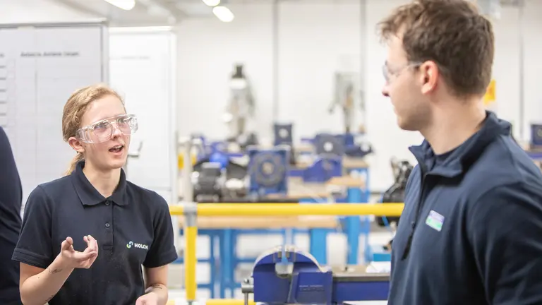 Two apprentices in blue shirts and safety goggles in a factory.