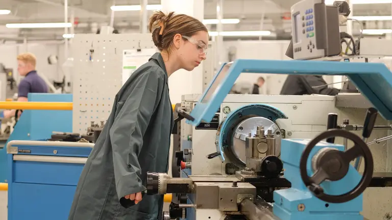 Woman wearing safety glasses and a lab coat adjusts a machine in a factory.
