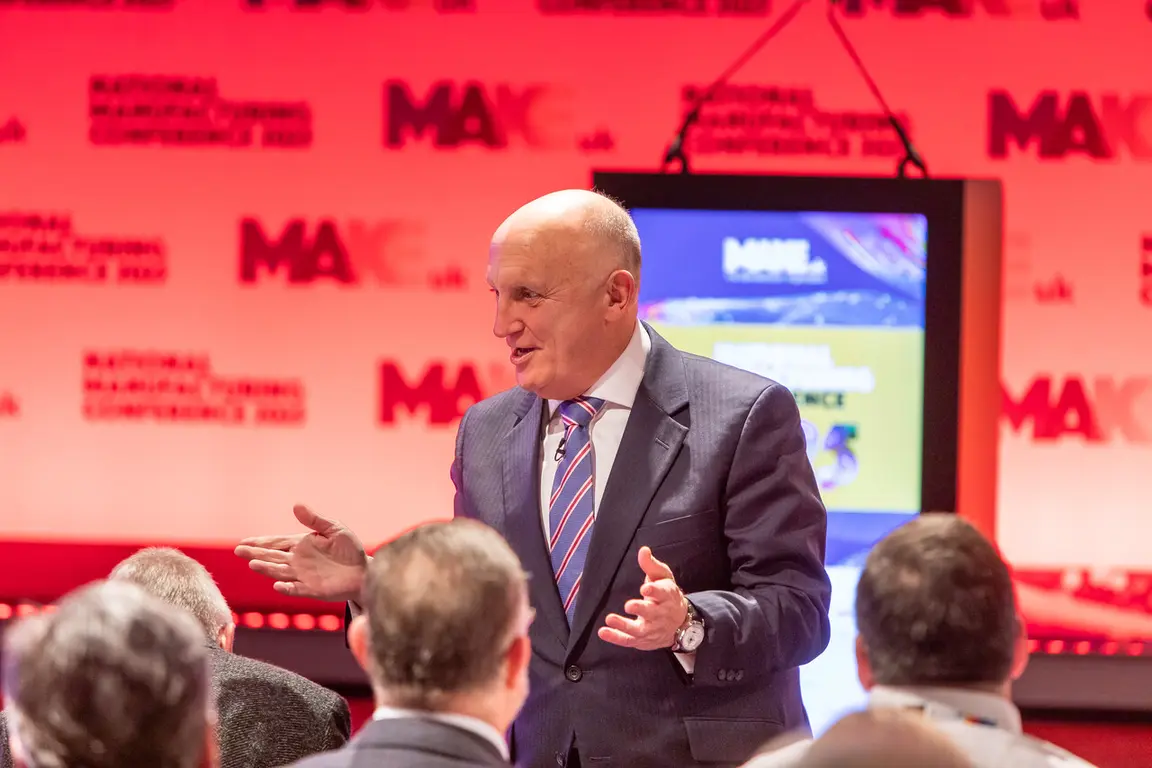 A man in a suit speaks at a conference while an audience listens.