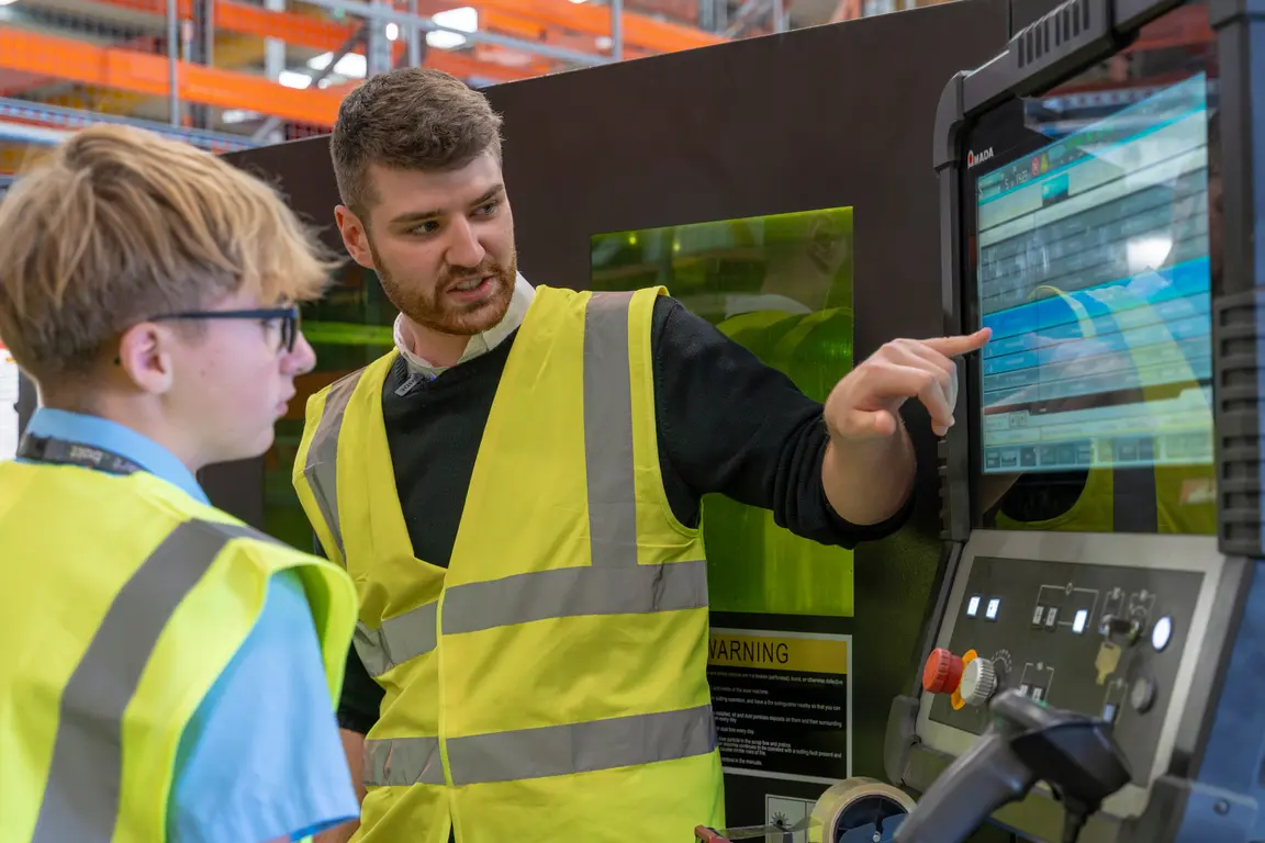 NMD Two men in safety vests standing in front of a large monitor.