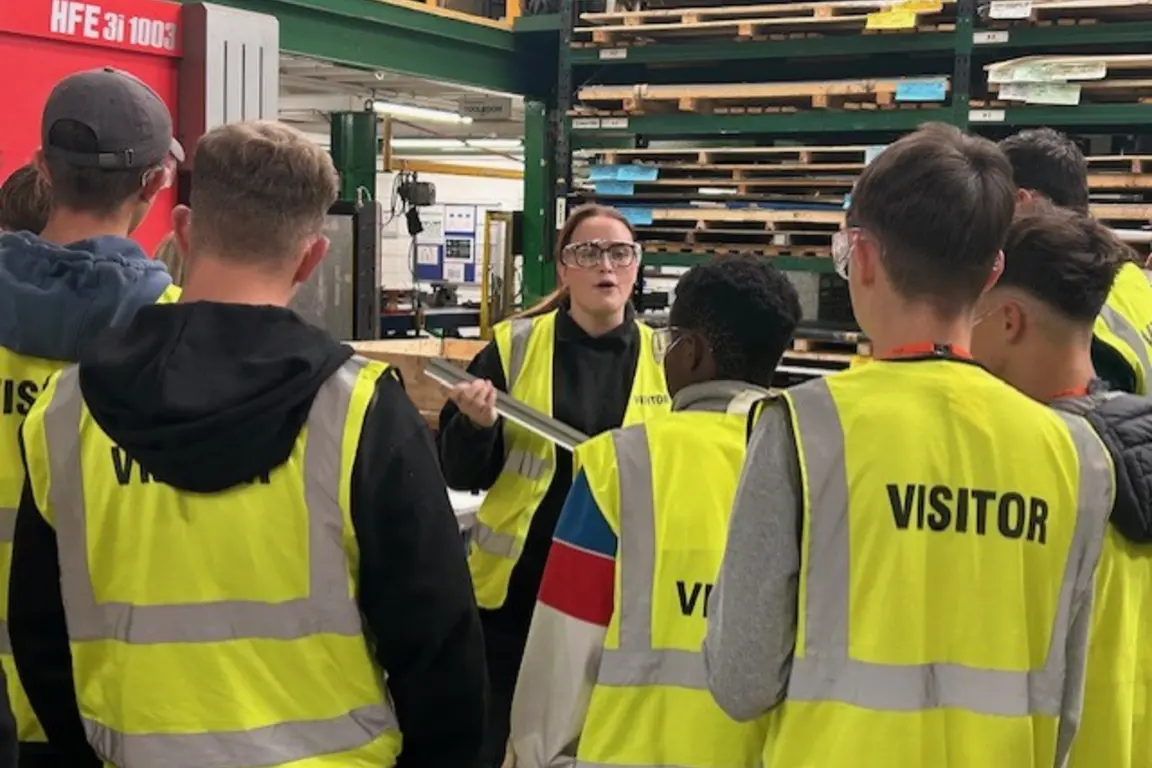 A group of NMD visitors in reflective yellow vests gather in a factory with a woman speaking.