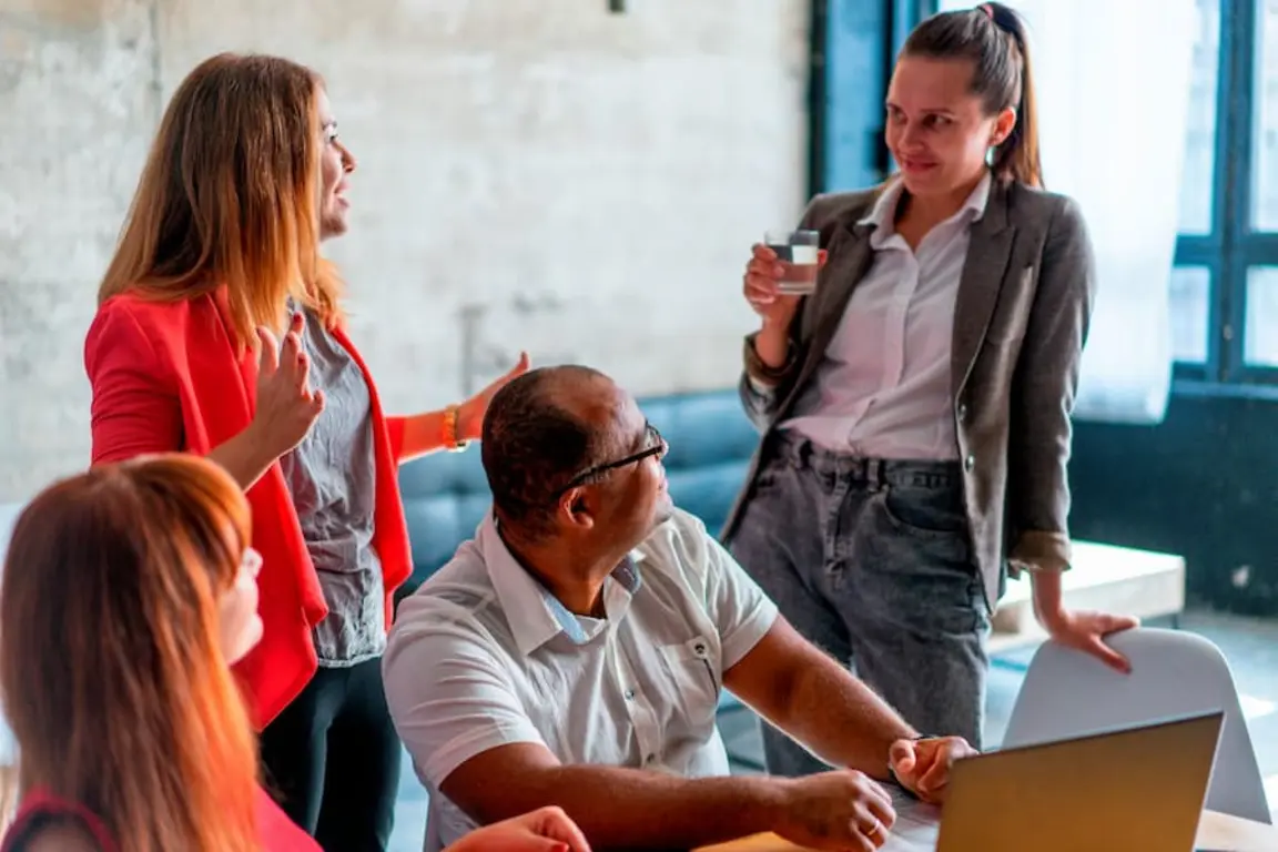 Four people in a room, one man sits with a laptop, three women talk.