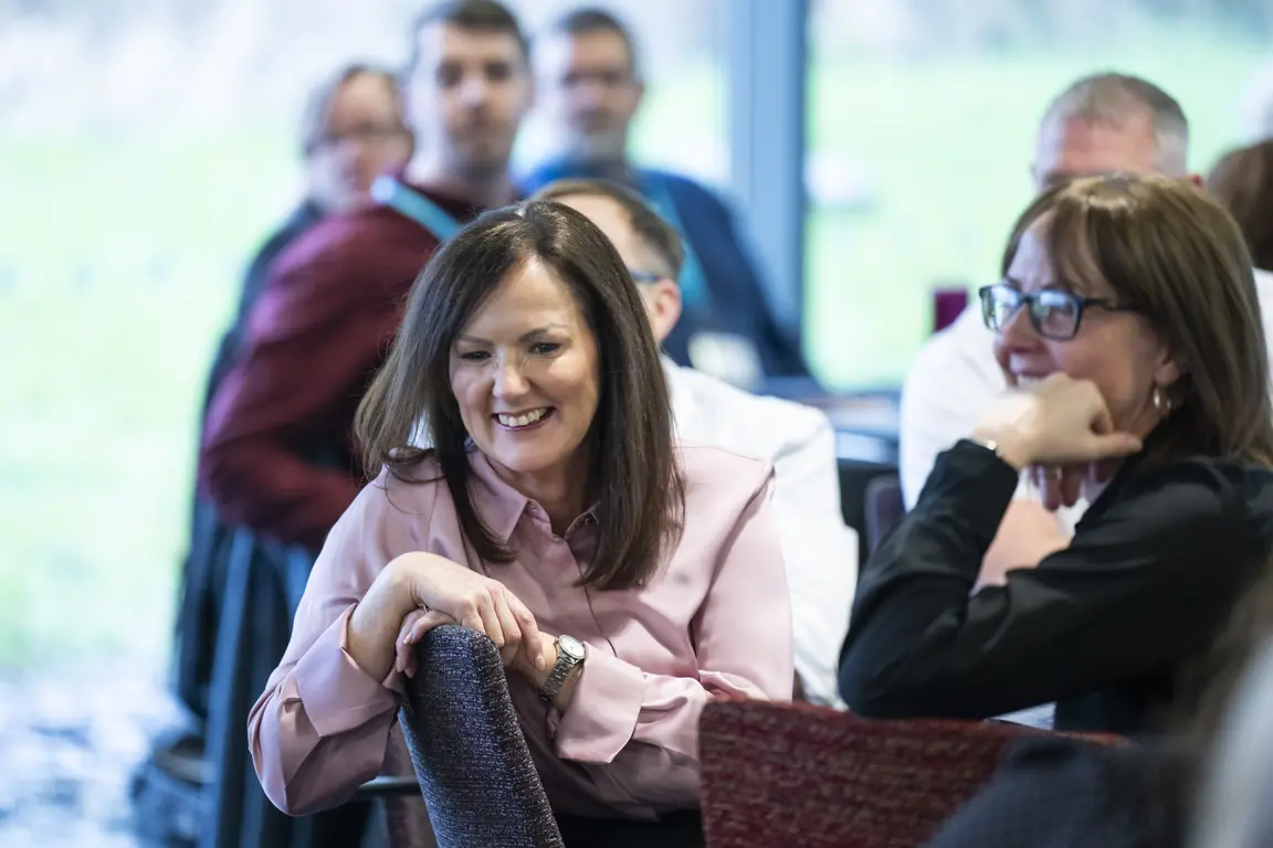 Woman smiling at a conference, with people sitting behind her, some blurry.