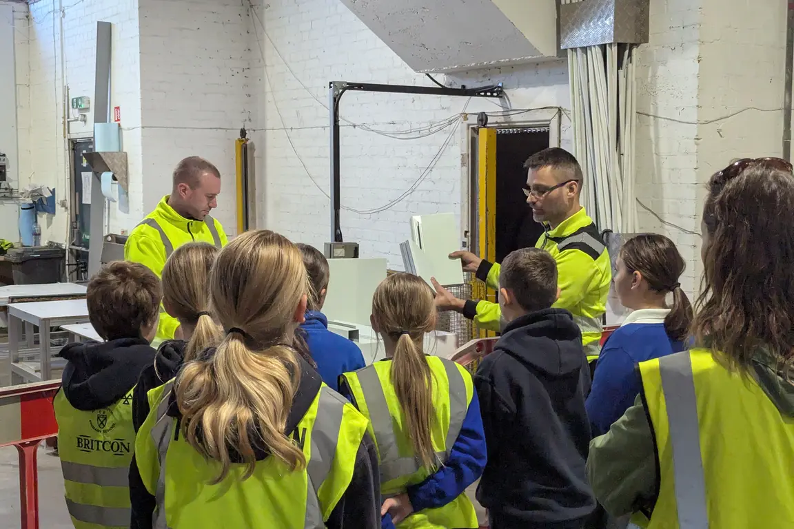 Children wearing yellow vests listen to a man explain something in a factory on national manufacturing day.