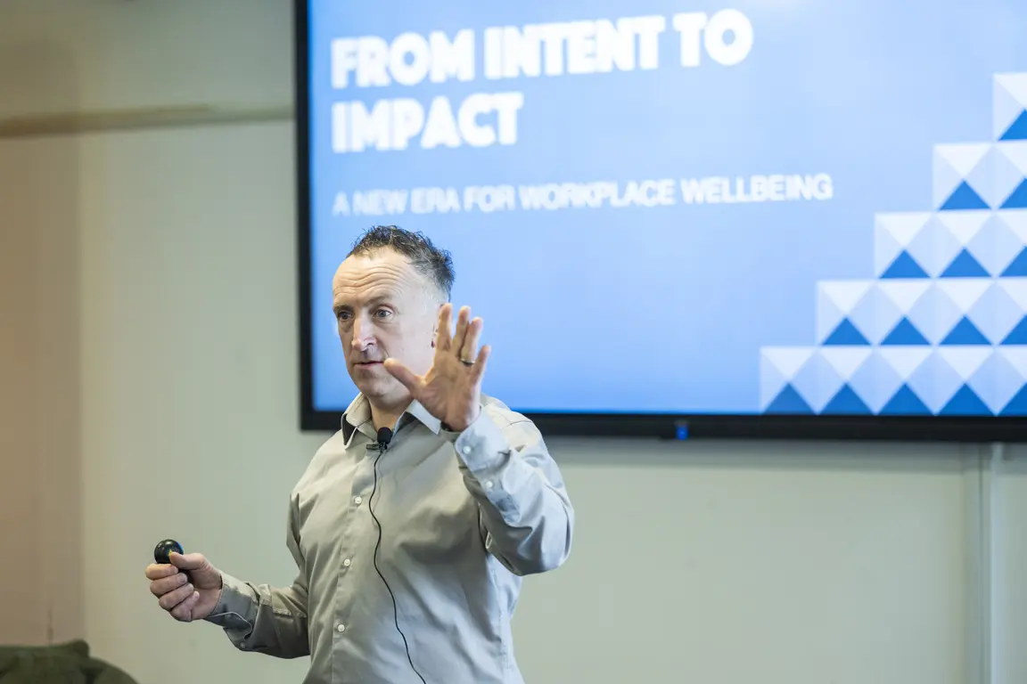 A man with short hair and a light-colored shirt gives a presentation in front of a screen that reads "From Intent to Impact" and "A New Era for Workplace Wellbeing".