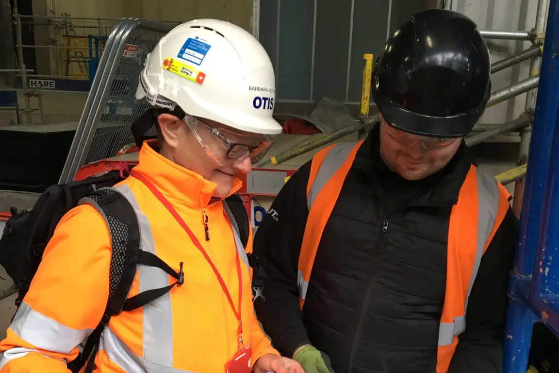 Two workers in orange vests and hard hats examining something in a warehouse.