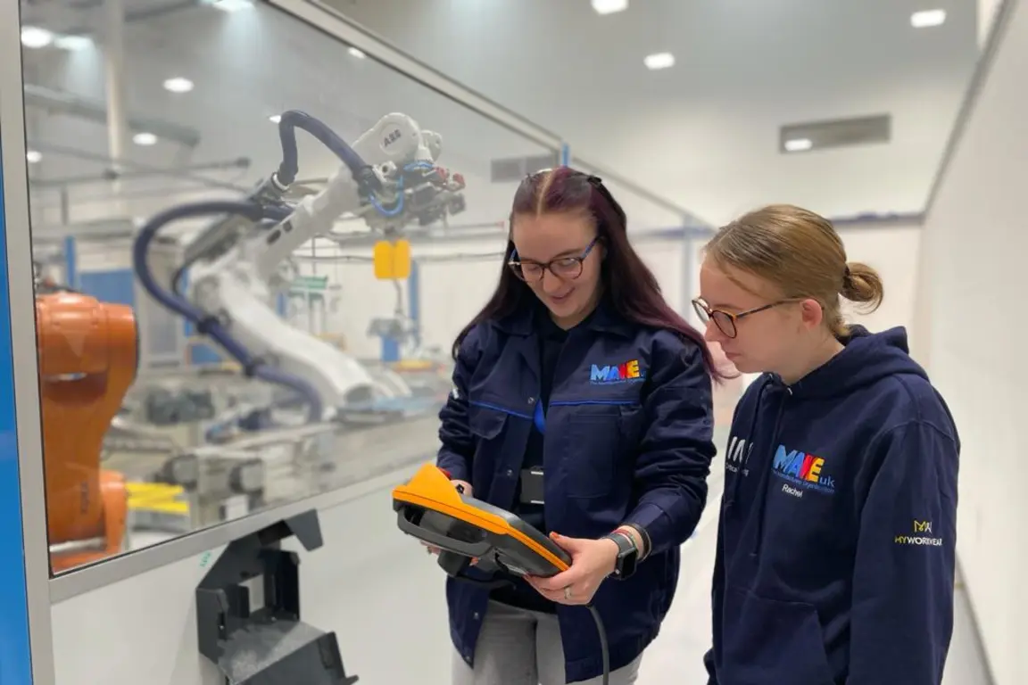 Two women examine a yellow tool inside an industrial building with glass walls and robotic arms.