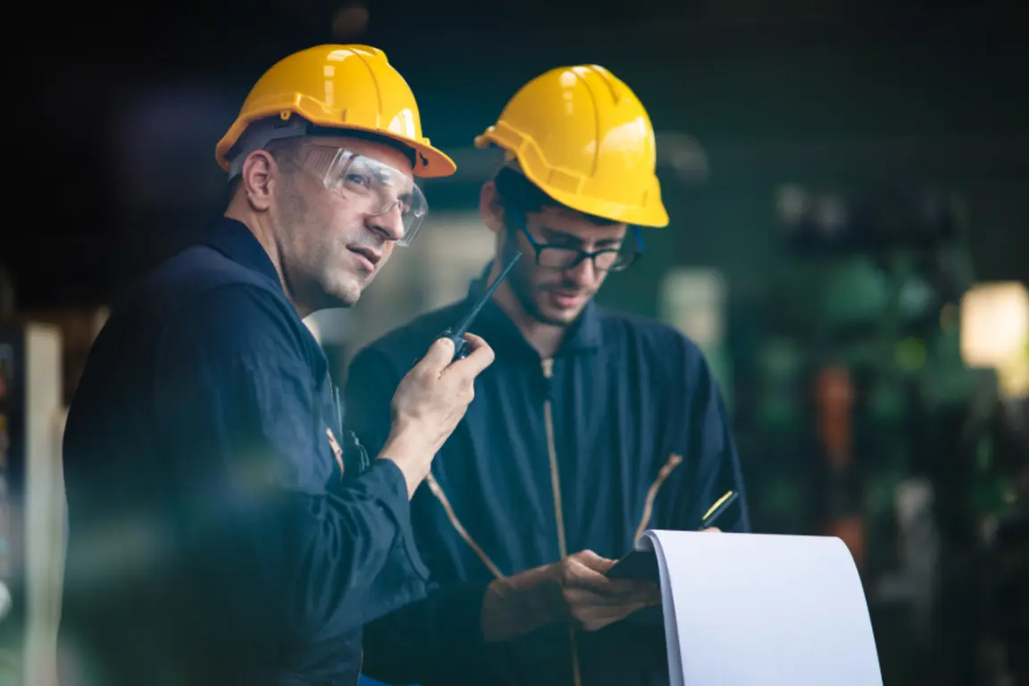 Two men in a factory, one holding a radio, the other looking at a phone.