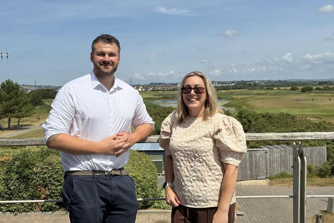 A man and woman stand on balcony against backdrop of green welsh landscape