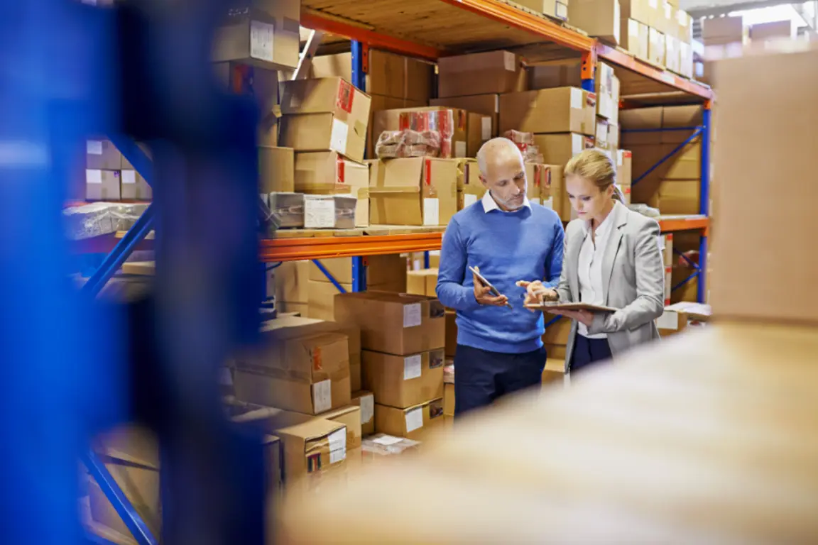 two business people in warehouse surrounded by shelves, boxes and stock examine a tablet