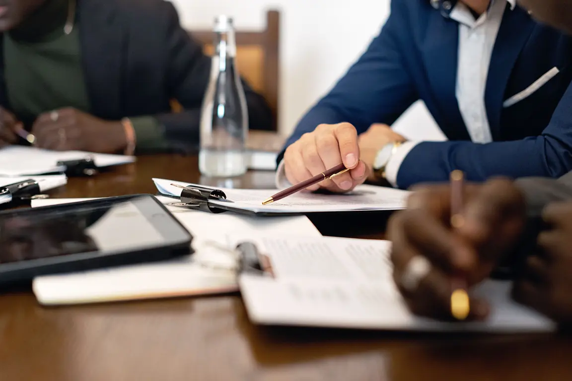 Three people in suits are writing at a table with a tablet, pens, and papers.