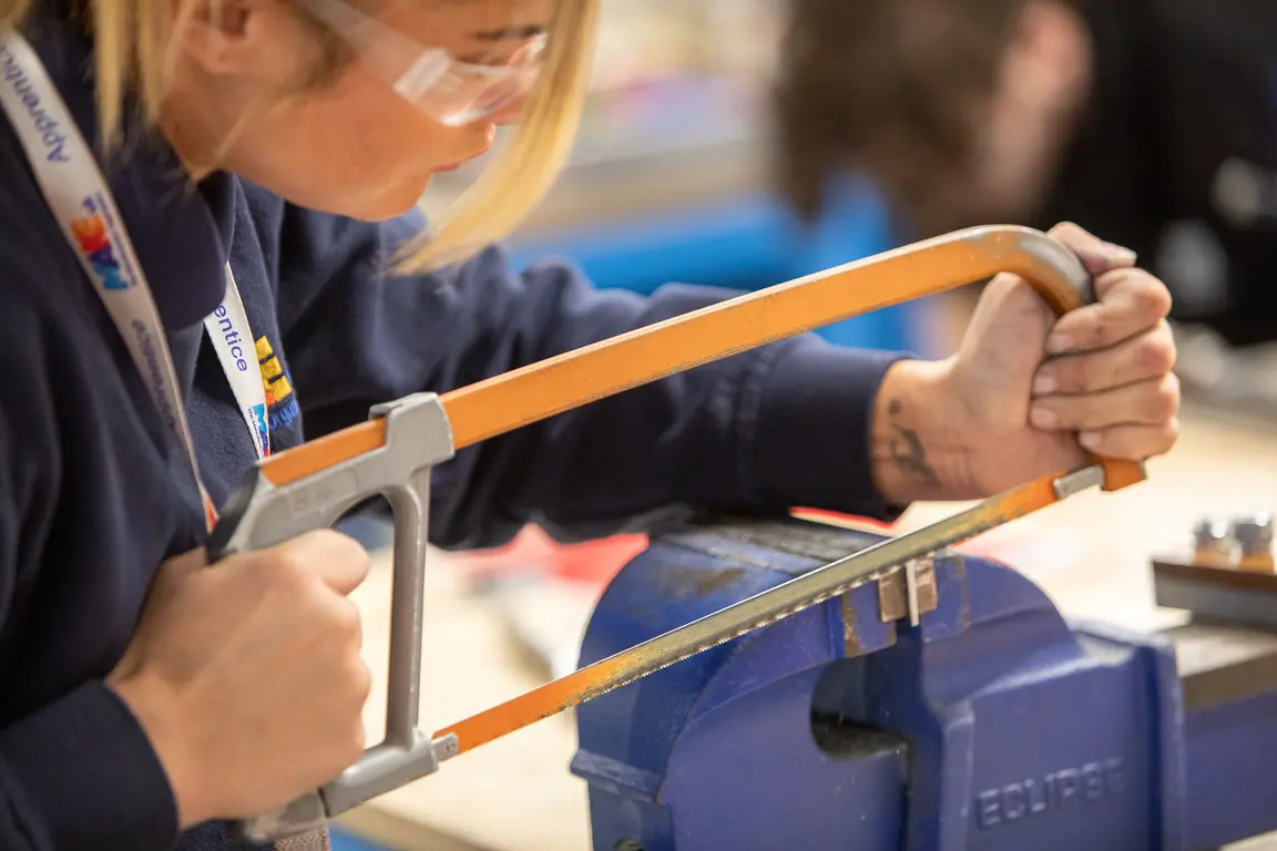Woman holding orange and gray hacksaw while using a blue metal clamp.