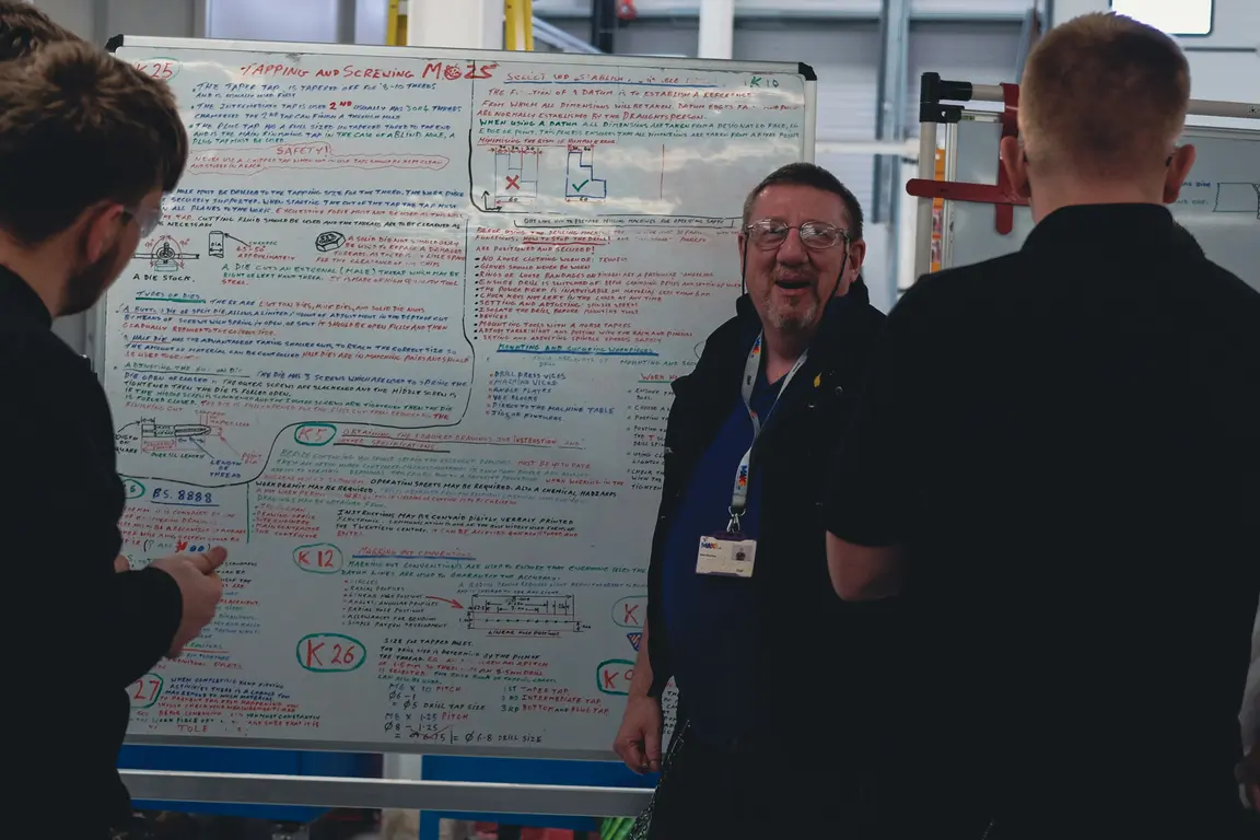 Four men in dark clothes stand in front of a large whiteboard filled with text and drawings.