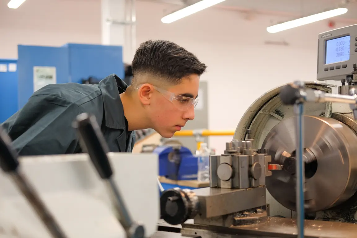 A young man in a workshop wearing safety goggles working on a large machine.